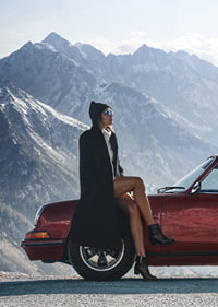 A stylish woman leans against a car with mountains in the background.-7