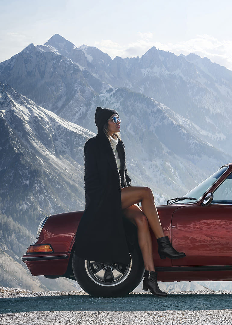 A stylish woman leans against a car with mountains in the background.-12