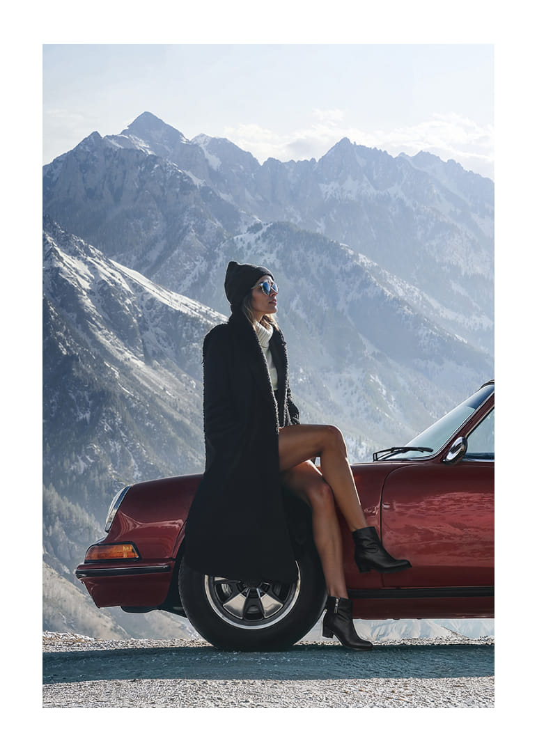 A stylish woman leans against a car with mountains in the background.-12