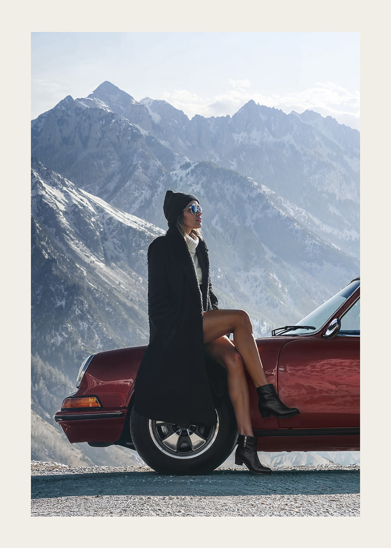 A stylish woman leans against a car with mountains in the background.-12
