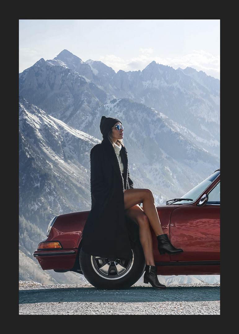 A stylish woman leans against a car with mountains in the background.-12