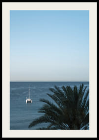A sailboat on the horizon on a calm sea with a palm tree in the foreground.-2