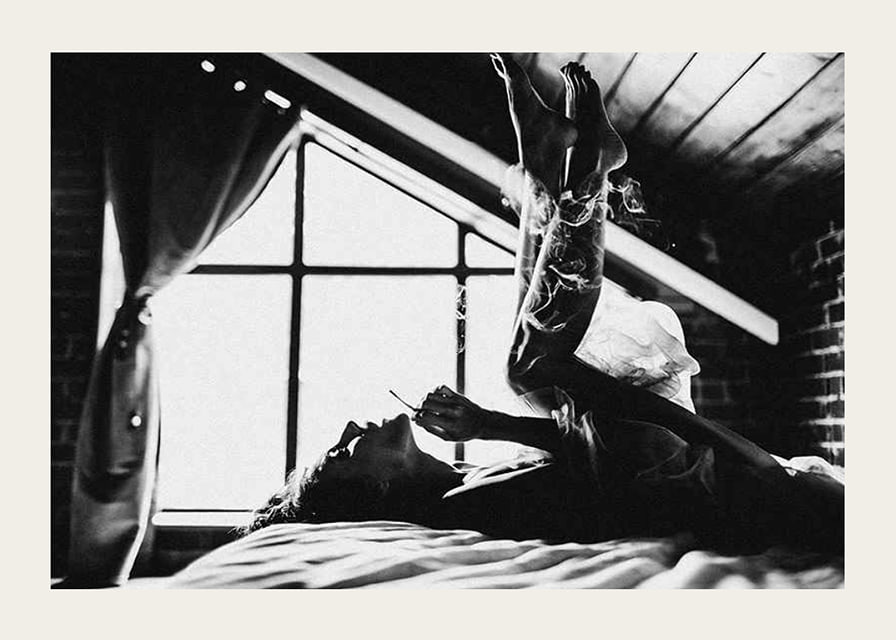 Black and white photograph of woman smoking in bed by window-12