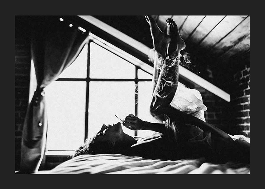 Black and white photograph of woman smoking in bed by window-12