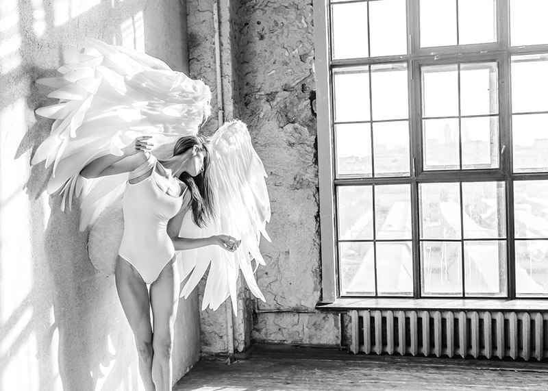 Black and white photo of woman with large angel wings by window-12