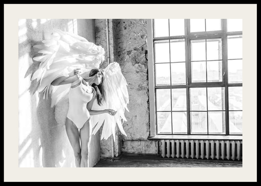Čiernobiela fotografia ženy s veľkými anjelskými krídlami pri okne-12