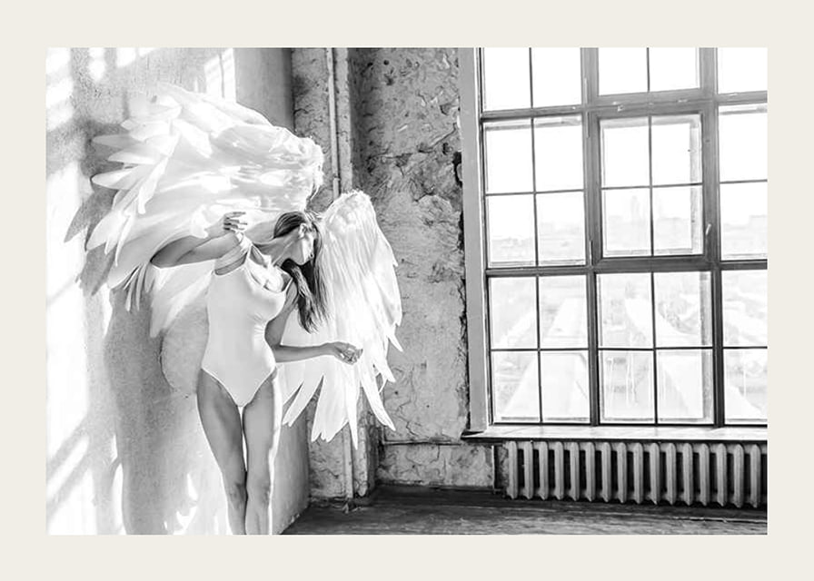 Black and white photo of woman with large angel wings by window-12
