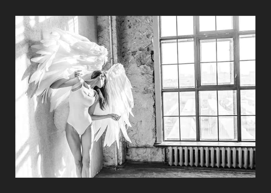 Čiernobiela fotografia ženy s veľkými anjelskými krídlami pri okne-12