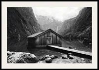 Black and white photo of wooden cabin by lake, surrounded by high mountains-2