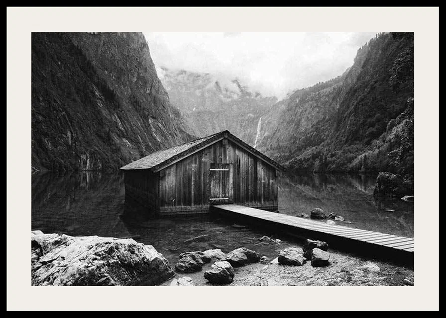 Black and white photo of wooden cabin by lake, surrounded by high mountains-12