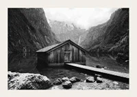 Black and white photo of wooden cabin by lake, surrounded by high mountains-3