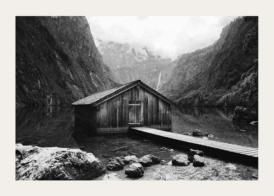 Black and white photo of wooden cabin by lake, surrounded by high mountains-12