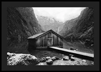 Black and white photo of wooden cabin by lake, surrounded by high mountains-4