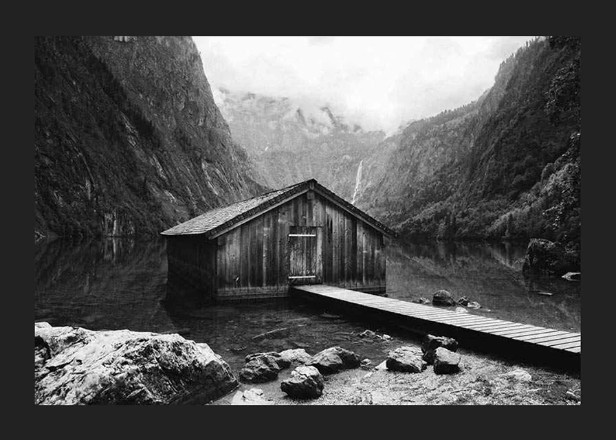 Black and white photo of wooden cabin by lake, surrounded by high mountains-12