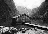 Black and white photo of wooden cabin by lake, surrounded by high mountains-7