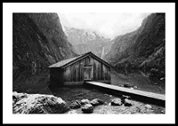 Black and white photo of wooden cabin by lake, surrounded by high mountains-0