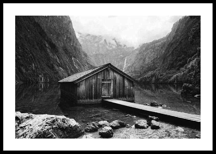Black and white photo of wooden cabin by lake, surrounded by high mountains-12