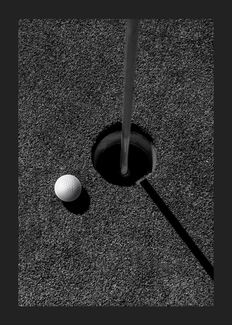 Black and white close-up of golf ball next to hole with flagpole-12