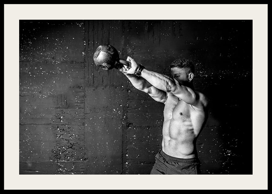 Black and white close-up of man exercising with kettlebell against raw concrete wall-12