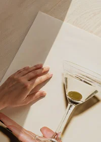 Photograph of martini glass with olive on hand against beige background-3