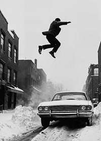 Black and white city scene with man jumping over snow-covered car-7
