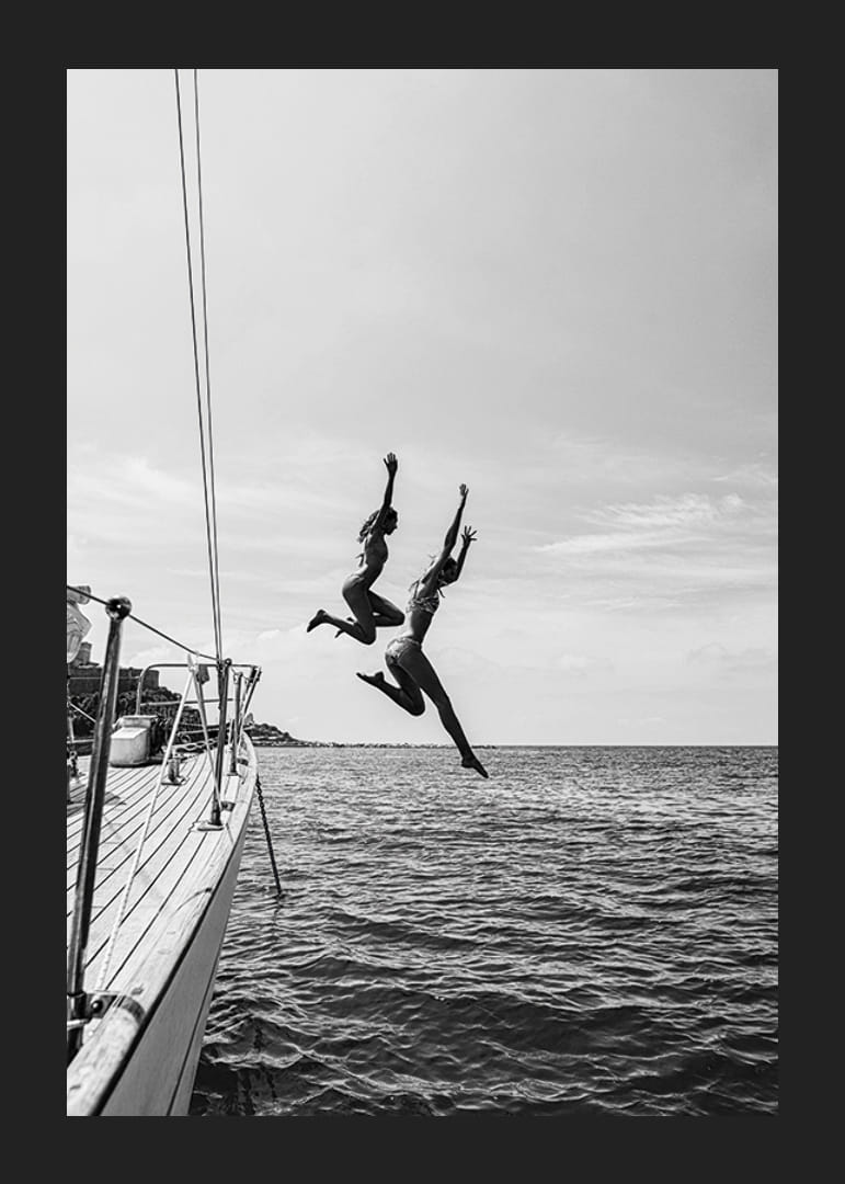 Svartvit poster med ett fotografi på två personer som hoppar från en segelbåt ner i havet-12